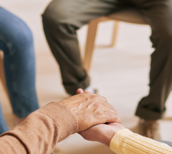 Cinco pessoas sentadas em cadeiras, em círculo, com as mãos dadas, expressando apoio, como em uma terapia em grupo.