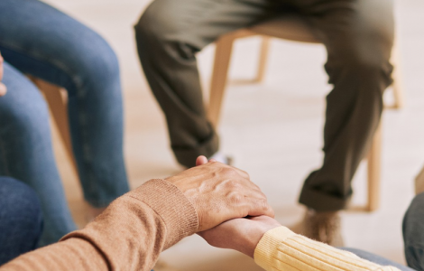 Cinco pessoas sentadas em cadeiras, em círculo, com as mãos dadas, expressando apoio, como em uma terapia em grupo.
