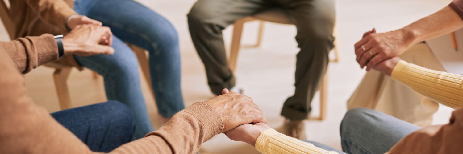 Cinco pessoas sentadas em cadeiras, em círculo, com as mãos dadas, expressando apoio, como em uma terapia em grupo.