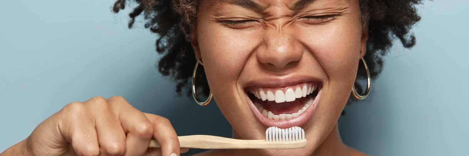Mulher sorrindo enquanto escova os dentes com escova de bambu, em frente a fundo azul claro.