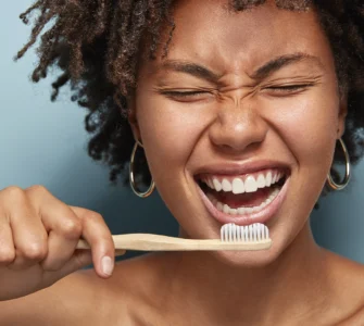 Mulher sorrindo enquanto escova os dentes com escova de bambu, em frente a fundo azul claro.