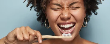 Mulher sorrindo enquanto escova os dentes com escova de bambu, em frente a fundo azul claro.