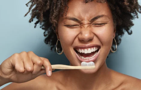 Mulher sorrindo enquanto escova os dentes com escova de bambu, em frente a fundo azul claro.