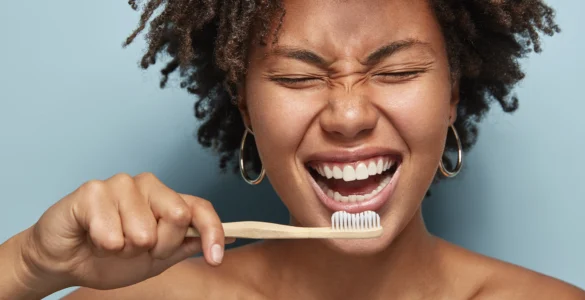 Mulher sorrindo enquanto escova os dentes com escova de bambu, em frente a fundo azul claro.