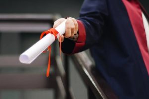 Pessoa vestindo traje tradicional de formatura, segurando um certificado de conclusão em papel, enrolado em um laço vermelho.