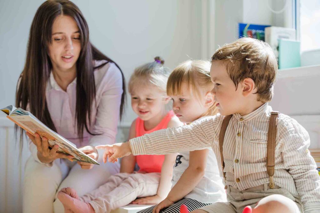 Um adulto acompanhado de três crianças lado a lado com um livro aberto e uma das crianças apontando para o interior do livro