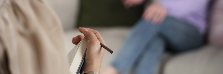 Imagem mostrando a mão de uma terapeuta fazendo anotações durante uma sessão de terapia, com um paciente ao fundo desfocado