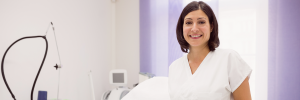 Profissional esteticista sorrindo, usando uniforme branco em ambiente clínico de trabalho.