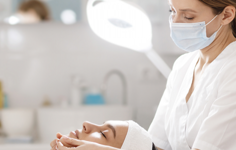 Profissional esteticista realizando tratamento na pele de uma cliente, deitada em sua frente, em ambiente clínico.