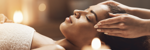 Mulher deitada em superfície branca, com os olhos fechados, enquanto recebe uma massagem estética profissional no rosto.