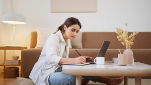 Mulher em uma sala, sentada em atividade de estudos, com um laptop aberto, escrevendo em um caderno e com uma xícara ao lado.