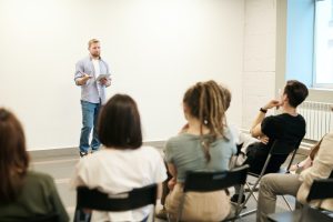 Ambiente de estudos e aprendizados no qual um professor fala ao público, composto por estudantes sentados e interessados.