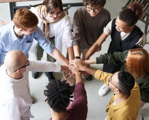 Na fotografia, há um grupo de pessoas multiétnico, composto em círculo, com as mãos unidas e sobrepostas, transmitindo uma simbologia de união e trabalho em equipe.