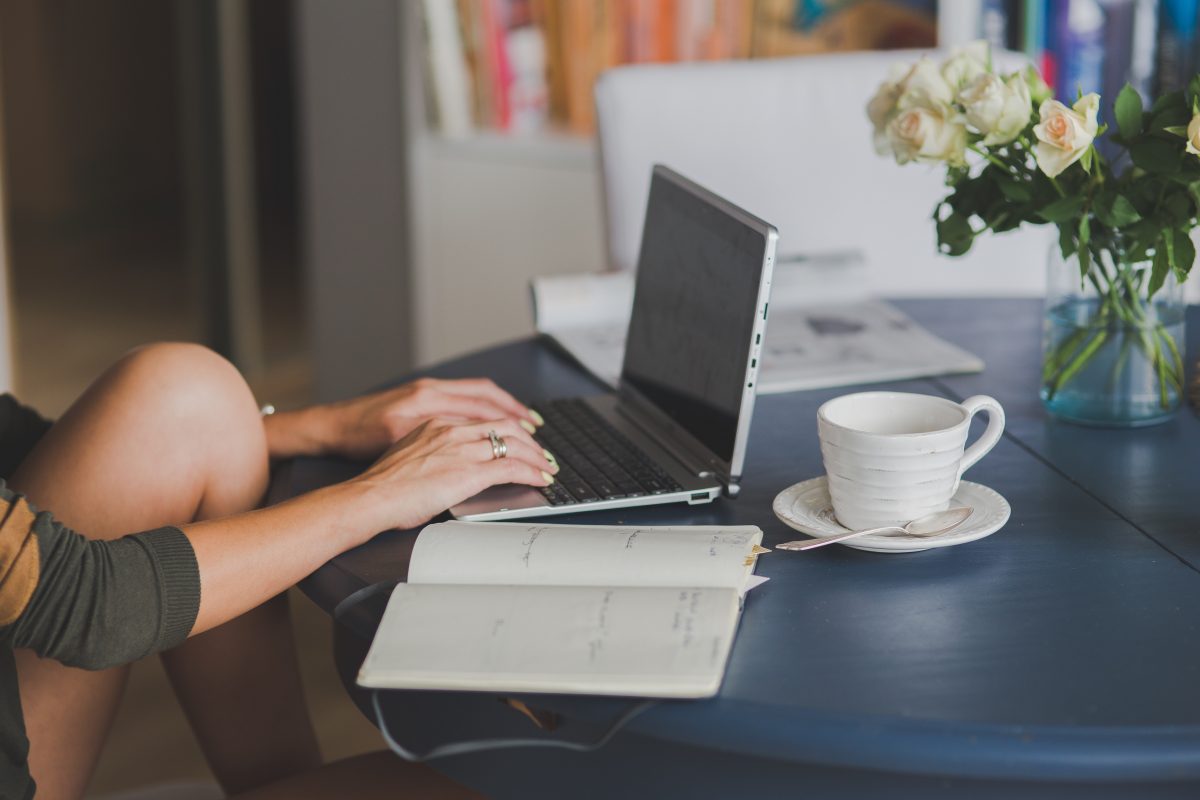 Mulher utilizando laptop em uma mesa de trabalho e estudos onde há uma xícara sobre um pires, flores, um livro e um caderno.