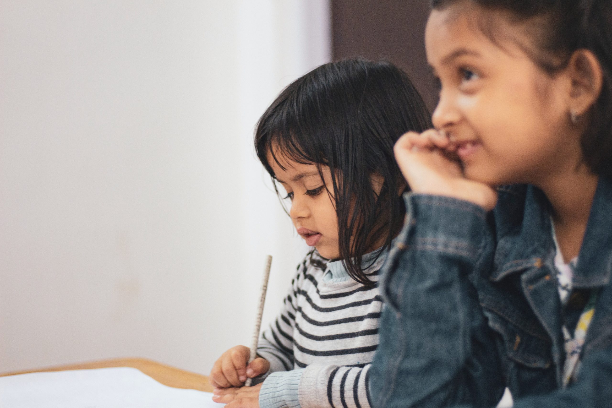 Duas crianças em ambiente de estudos posicionadas perante uma mesa. Uma delas escreve em uma folha de papel, a outra escuta.