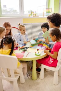 Professoras ensinam crianças sentadas, ao redor de uma mesa redonda, auxiliando-as em atividade educacional lúdica e criativa.