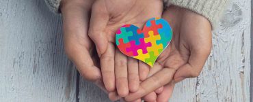 Quatro mãos, sobrepostas uma à outra, seguram o símbolo em formato de coração representativo do diagnóstico de autismo.