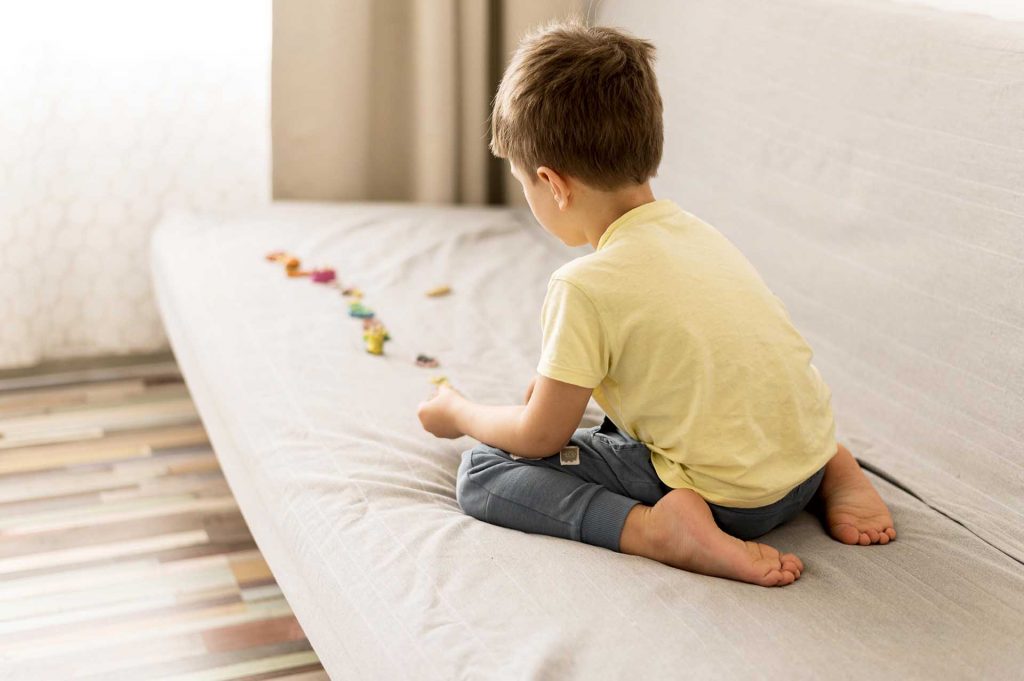 Criança sentada em um sofá cinza, brincando e alinhando pequenos brinquedos coloridos
