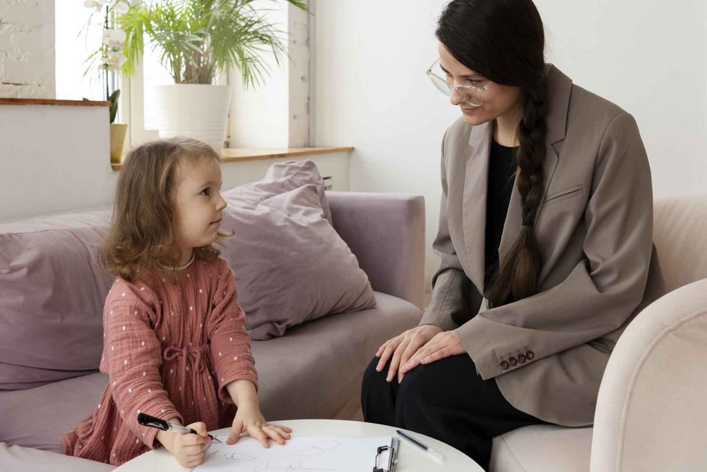 Criança desenhando em uma mesa pequena enquanto conversa com uma psicóloga em um ambiente aconchegante com plantas ao fundo.
