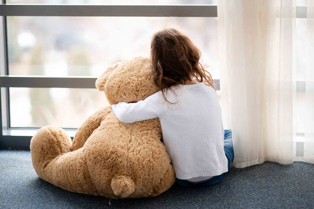 Criança sentada no chão, abraçando um urso de pelúcia grande, olhando pela janela