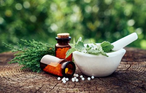 Em uma mesa de madeiras, dois frascos de medicamento fitoterápico estão ao lado de um recipiente com folhas, um deles caído.