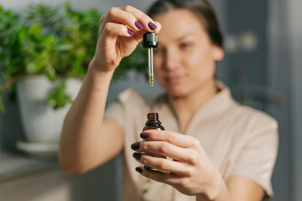 Mulher segurando um conta-gotas em uma mão e um frasco de medicamento na outra, com plantas desfocadas ao fundo