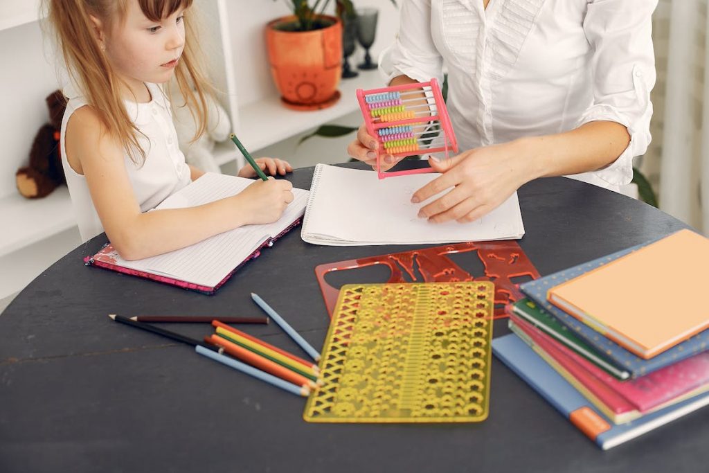 Criança estudando com um adulto, usando um ábaco e cercada por cadernos, lápis de cor e réguas geométricas