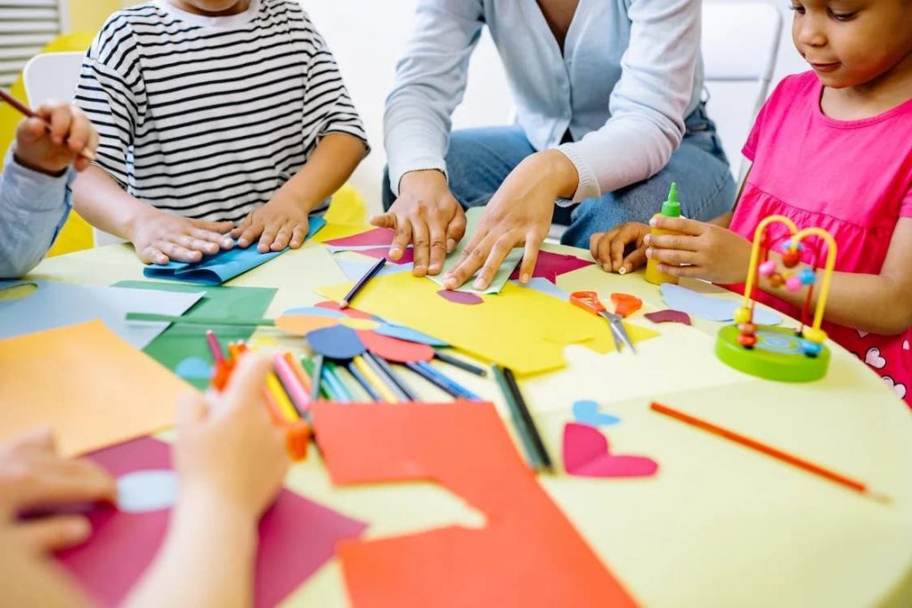 Crianças fazendo atividades de arte em uma mesa redonda, utilizando papel colorido, tesouras, cola e lápis de cor, acompanhadas por um adulto.