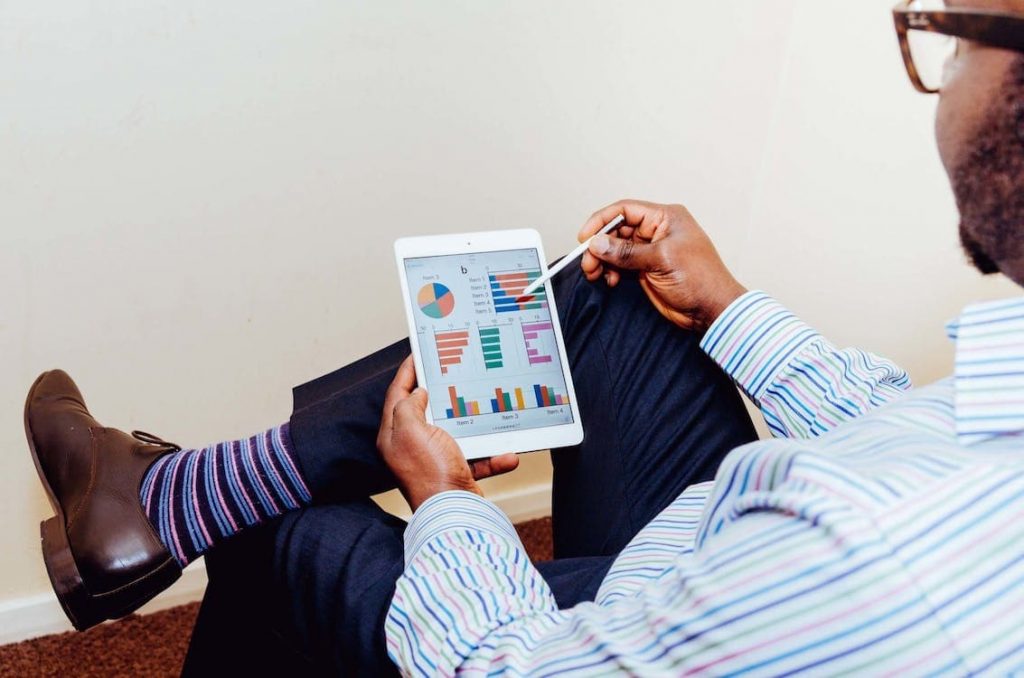 Homem analisando gráficos e planilhas em um tablet, utilizando uma caneta stylus, vestido com roupas formais e meias coloridas