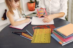 Criança do sexo feminino observando instruções de sua Psicopedagoga enquanto segura um lápis e um caderno para anotações.