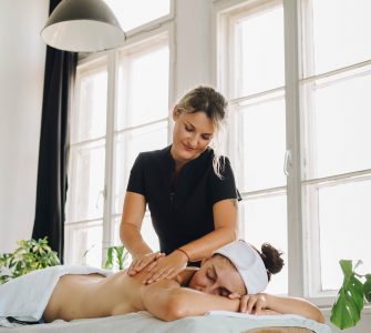 Profissional realizando massoterapia em paciente, a qual está deitada em de bruços, recebendo os cuidados de saúde.