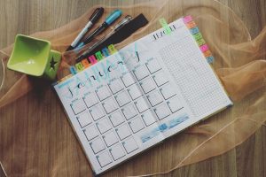 Agenda disposta sobre a mesa, demonstrando calendário do mês de janeiro, ao lado de canetas coloridas e um objeto decorativo.