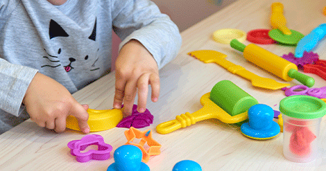 Criança de roupa cinza brincando em uma mesa utilizando diversos materiais coloridos