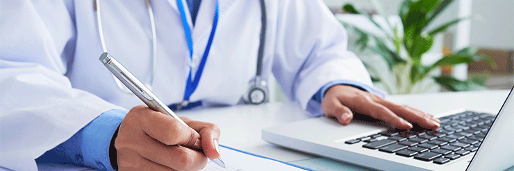 Médico escreve em um bloco de notas enquanto usa um laptop, vestindo jaleco branco e estetoscópio no pescoço