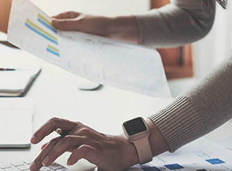 Pessoa analisa documentos financeiros em frente ao laptop, com smartwatch no pulso e gráficos na mesa