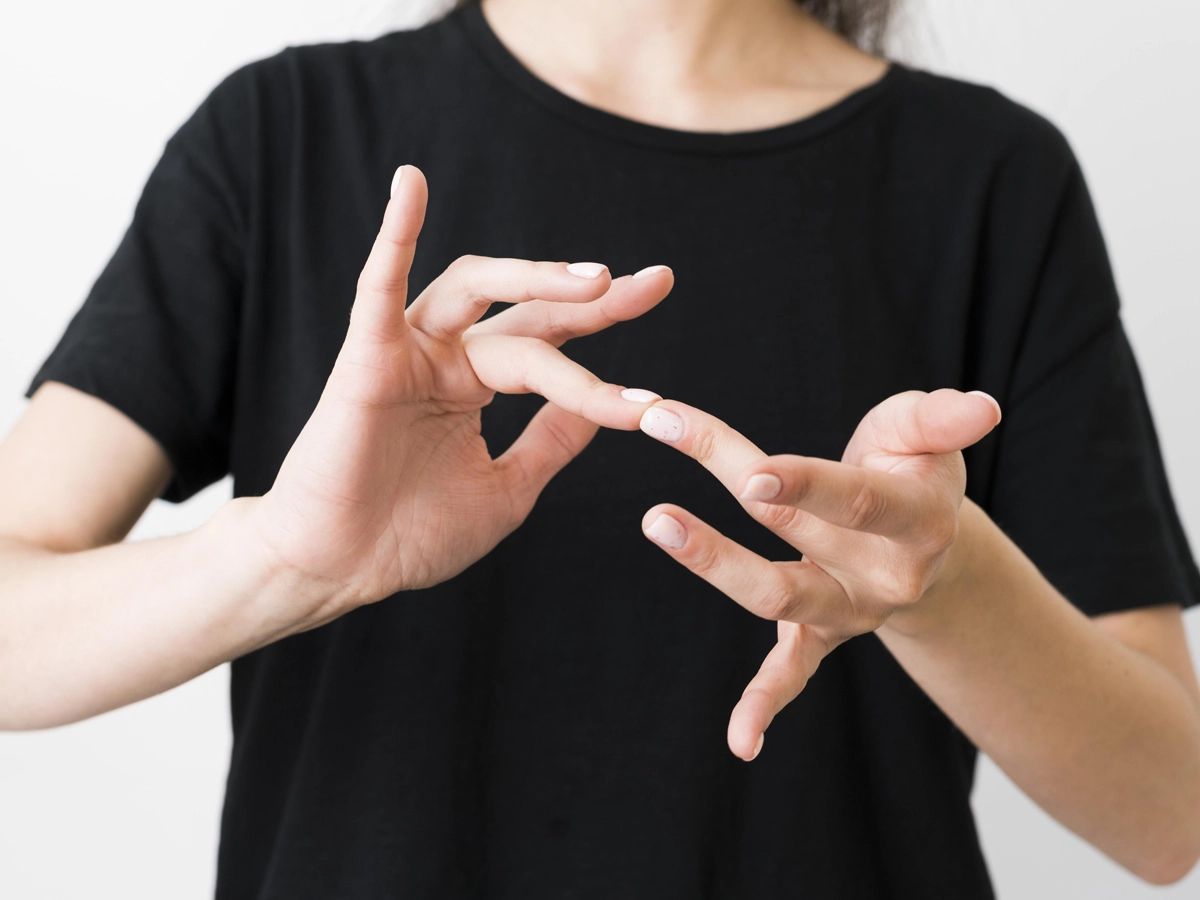 Em um fundo branco, uma pessoa usando camiseta preta utiliza as mãos para se comunicar em libras.