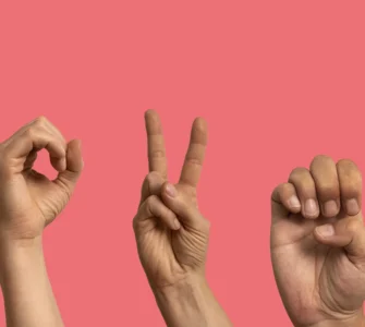 Imagem mostrando três mãos fazendo gestos em Libras (Língua Brasileira de Sinais) com um fundo rosa.