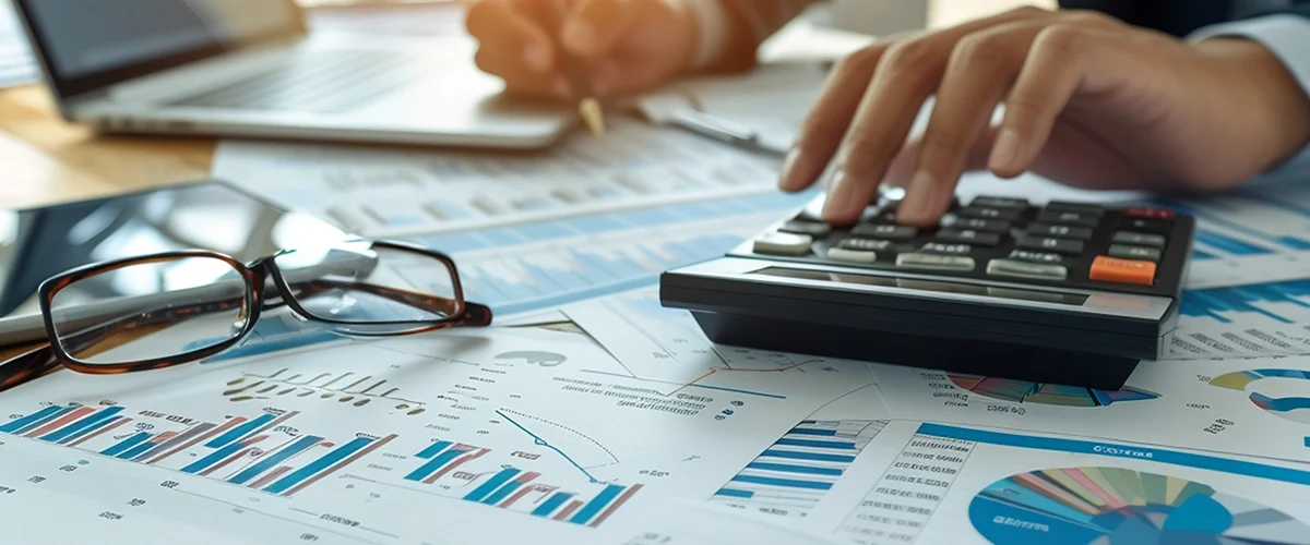 Pessoa utilizando calculadora e analisando gráficos e relatórios financeiros sobre uma mesa com laptop e óculos.
