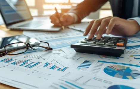 Pessoa utilizando calculadora e analisando gráficos e relatórios financeiros sobre uma mesa com laptop e óculos.