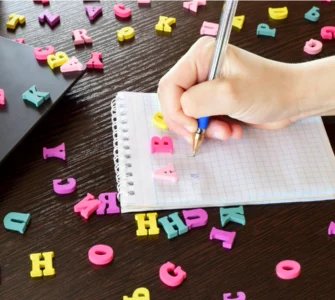Em uma mesa de madeira escura, uma mão escreve em uma caderneta branca, cercada de formas de letras do alfabeto coloridas.