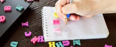 Em uma mesa de madeira escura, uma mão escreve em uma caderneta branca, cercada de formas de letras do alfabeto coloridas.