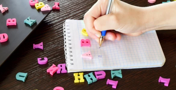 Em uma mesa de madeira escura, uma mão escreve em uma caderneta branca, cercada de formas de letras do alfabeto coloridas.