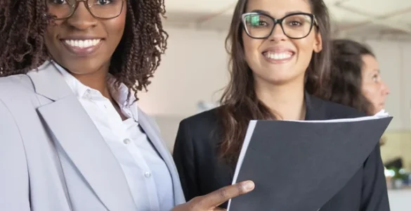 Grupo de mulheres profissionais sorrindo em um ambiente corporativo, segurando documentos e conversando.