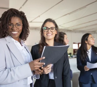 Grupo de mulheres profissionais sorrindo em um ambiente corporativo, segurando documentos e conversando.