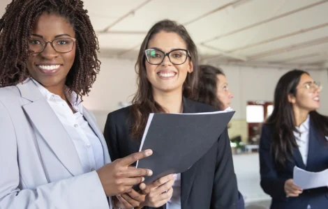 Grupo de mulheres profissionais sorrindo em um ambiente corporativo, segurando documentos e conversando.
