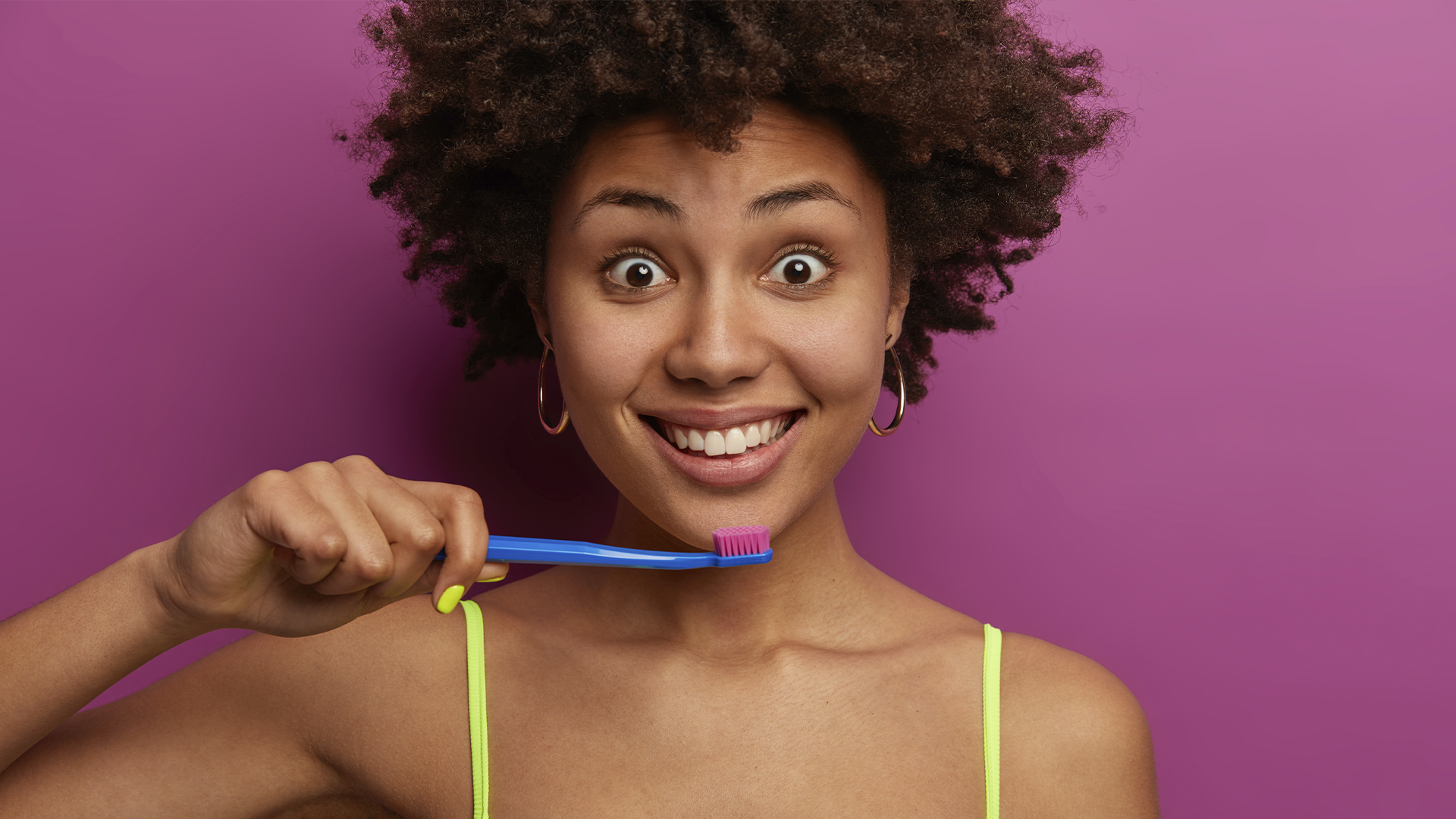 Mulher sorridente segura escova de dentes azul com cerdas rosas diante de fundo rosa vibrante.