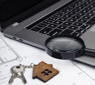 Chaveiro de casa, chaves, lupa e laptop sobre planta arquitetônica, representando tecnologia no setor imobiliário.