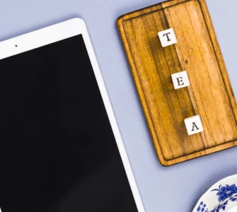 Tablet branco, tabuleiro de madeira com letras formando "TEA" e pratos de porcelana azul sobre fundo lilás.