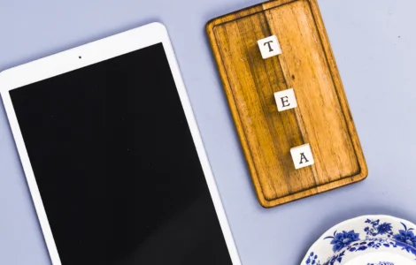Tablet branco, tabuleiro de madeira com letras formando "TEA" e pratos de porcelana azul sobre fundo lilás.