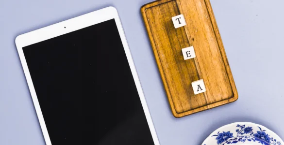Tablet branco, tabuleiro de madeira com letras formando "TEA" e pratos de porcelana azul sobre fundo lilás.
